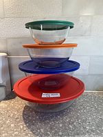 Four clear Pyrex round glass mixing bowls stacked nested, each with a different color plastic lid: green, orange, blue, red from smallest to largest.