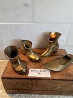 Four vintage brass shoe figurines displayed on a wooden box, showing various styles including heels, boots, and flats.