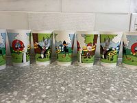 Eight vintage Looney Tunes cups displayed in a row showing colorful character artwork including Bugs Bunny, Daffy Duck, and others in outdoor scenes.