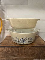 Stacked view of two vintage Pyrex casserole dishes, beige speckled with blue folk art floral pattern on the smaller dish.