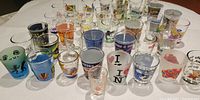 Photo showing a large assortment of 42 shot glasses arranged on a table with various designs and colors visible