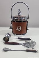 Front angle of equestrian themed ice bucket with horse head lid handle, brown leather wrap and horseshoe decor, accompanied by four matching bar tools with wooden handles.