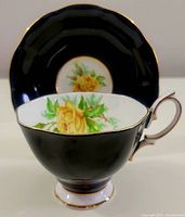 Tea cup and saucer set with black exterior and yellow rose floral decoration on the interior of the cup and center of the saucer. Gold trim highlights edges and handle.