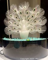 Lamp shown with fiber optic peacock tail unlit against dark background, clear fiber optic strands with painted eye patterns on tail feathers, frosted peacock figure, black round base with glass gems around.