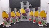 Photo showing the frosted glass pitcher surrounded by six clear drinking glasses with fruit patterns.