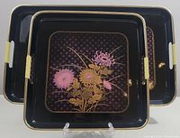 Two black lacquer rectangular serving trays with pink, purple and gold floral decoration and cream handles stacked nested.