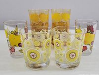 Photo showing all six glasses arranged in two rows: two tall gold floral Libbey glasses at the back center, two medium fruit pattern Morning Star glasses on each side, and two short psychedelic pattern Ikea glasses in front center.