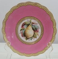 Front view of vintage ceramic pedestal cake plate showing scalloped pink rim, gold decorative border and central pear illustration.
