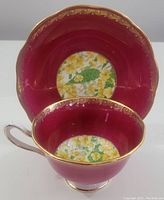 Tea cup and saucer displayed with saucer behind cup showing burgundy color, gold ornate edge design, and floral pattern in center