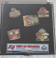 Top view photo showing all six enamel pins mounted on backing card with black background. Pins depict Buccaneers Super Bowl history and scores. Includes limited edition card numbered 3462 of 5000.