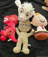 Four assorted plush toys including red dinosaur, white goat, mummy, and a cartoon character with closed eyes in suit and tie.