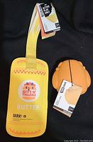 Photo shows two new dog toys with original tags: a yellow rectangular toy labeled 'Give N' Thanks Butter Bark' with a handle, and an orange paw-shaped textured toy.