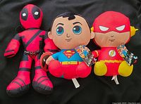 Three plush toys of Deadpool, Superman, and The Flash placed side by side on black surface.