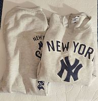 Front view of folded light grey New York Yankees sweatshirt with large embroidered logo and text in navy blue and white, paired with matching sweatpants folded beside it