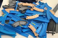 Various blue plastic curved and straight Thomas The Train track pieces and tan wooden track pieces piled together