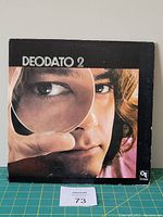 Front cover of Deodato 2 vinyl LP showing close-up photo of a man's face with a magnifying glass, with some wear and edge damage visible.