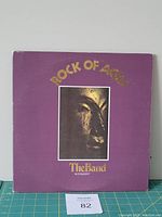 Front of the 'Rock of Ages' vinyl LP by The Band showing purple gatefold cover with gold title and image of sculptural face.