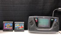 Sega Game Gear handheld console with screen visible, two cartridges positioned beside it