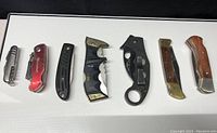 Seven folding pocket knives displayed side by side showing diverse handles and design.