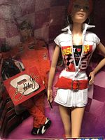Close-up of Barbie doll dressed in NASCAR-themed white and red racing outfit standing next to Dale Earnhardt Jr. figure holding signed flag inside box.