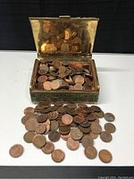 Open rectangular copper box filled and overflowing with copper pennies on white surface with black background.
