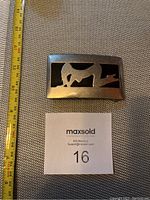 Top view of a rectangular silver-tone metal belt buckle with a cut-out horse design and black background, next to measuring tape showing about 3.5 inches width.