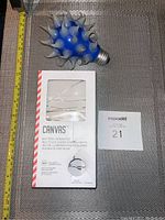 Blue decorative bulb with white spike design and box of Canvas battery-operated LED string lights on a textured surface with measuring tape showing approximate size.