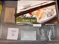 Overview of sushi set components including chopsticks, ceramic plates, spoon, and packaging box, all on a textured surface.