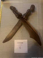 Two wooden African decor figurines with blade bases, viewed top down and close-up showing carved human figures seated on each blade.