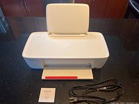 Front view of white HP DeskJet 1112 printer with paper tray open, power cable and USB cable placed in front.