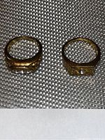 Photo of two gold toned men's rings placed side by side on a textured background showing top and band detail.