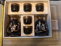 Full view of the sake set showing 2 black decanters with white script and 5 matching cups in a white box