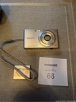 Sony Cyber-shot DSC-W320 silver digital camera with wrist strap and rechargeable battery placed beside it on a gray fabric surface.