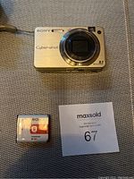 Silver Sony Cyber-shot DSC-W150 digital camera top/front view showing lens, flash, branding, and specifications with Sony NP-BG1 battery pack and auction ID card.