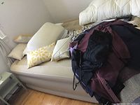Photo showing an inflated cream/light beige airbed with bedding including pillows and blankets piled on top, located near a window and wooden floor.