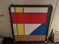 Framed acrylic painting showing geometric shapes in red, yellow, white, blue, and a brick pattern block at bottom, with decorative black frame