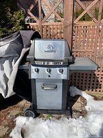 Front view of the Broil King Monarch BBQ Grill with three control knobs, metal body, and side shelf on the right. The grill is positioned outdoors on stone ground with snow around.