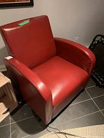 Red bonded leather armchair with cushioned seat and backrest, rounded armrests shown from front and side angles on dark tiled floor near wall with electrical outlet.