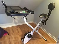 Side angle of white FitDesk exercise bike showing frame, seat with backrest, pedals, and desktop console.