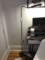 Black adjustable floor LED lamp standing beside a desk. Cord plugged in with inline switch visible. Lamp head is elongated rectangular shape.