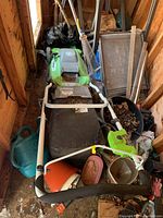 Greenworks electric lawn mower stored indoors with various gardening items around, showing the entire mower assembled with collection bag and handlebar.
