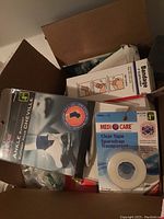 Photo showing ankle support and clear transparent tape in packaging, with part of a bandages box and other first aid kit materials visible inside a cardboard box.