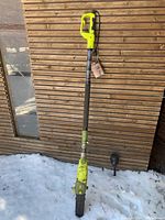 Full length view of green and black Sun Joe pole chainsaw standing upright in snow against wood backdrop.