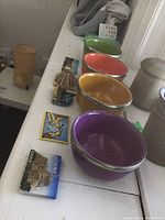 Side view showing four colorful bowls in purple, yellow, orange, and green, fridge magnets lined up, and pottery containers.