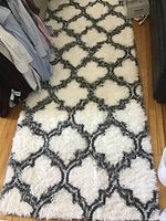 Full view of the white plush area rug with black geometric trellis pattern placed on wood floor beside clothing and furniture.