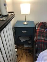 Side table positioned against wall with metal base lamp illuminated on top, showing marks and wear on table surface and drawer.