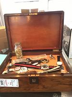 Open wooden box displaying multiple wristwatches with metal and leather straps, a glass jar, and loose clasps inside.