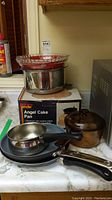 Photo showing Angel Cake Pan in original box, two frying pans with black handles (one T-Fal), glass pot with lid, and stacked glass pie dishes on countertop.