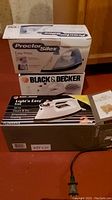 Photo shows three irons: boxed Proctor Silex Easy Press model 17400, boxed Black and Decker Light'n Easy Iron FL293WHS, and the Black and Decker X320 iron out of the box, all arranged upright.