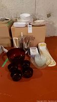 Wide view of kitchenware and dinnerware including glass bowls, coffee percolator, plates stacked in two piles, and plastic dish drainer on a basement floor.